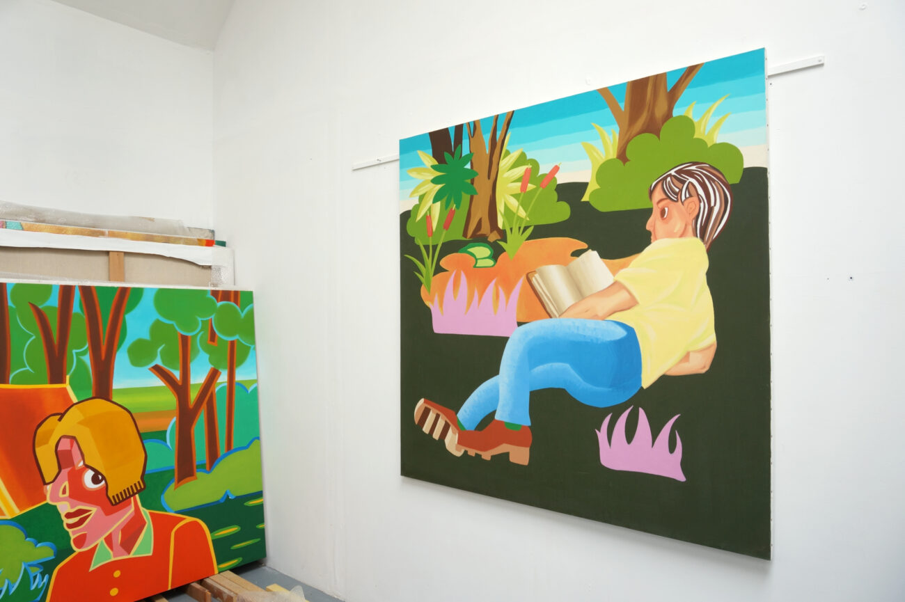 Peinture accrochée au mur dans l'atelier de l'artiste peintre Benoît Andro qui représente un personnage en train de lire un livre en plein-air. A gauche une autre peinture partiellement dissimulée.